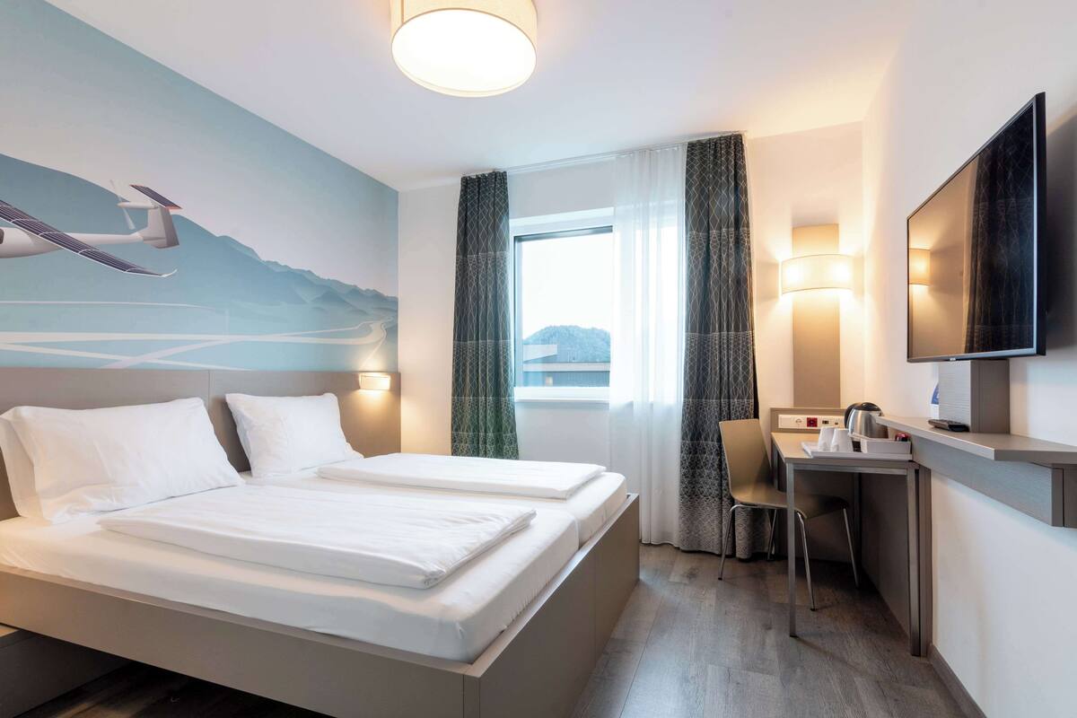 A hotel room with a bed, a desk, a chair, a TV, and a window with curtains.