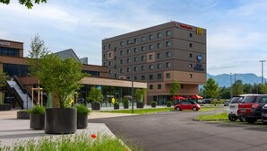 A hotel with a modern design, featuring a large building with multiple floors and a flat roof. There are outdoor seating areas with umbrellas, and a parking lot with several cars.