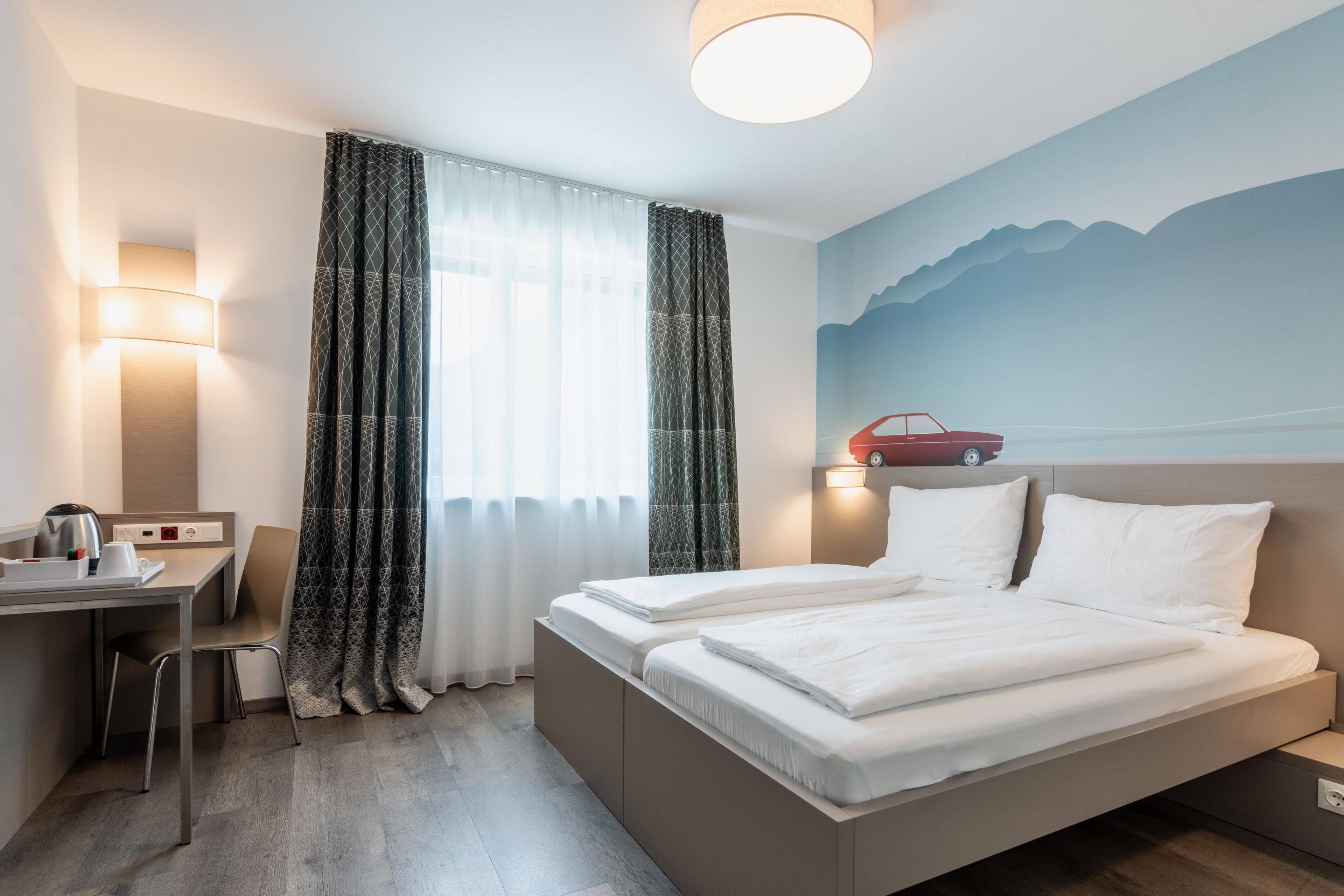 A hotel room with a bed, a desk, a chair, a coffee maker, and a wall with a mountain painting.