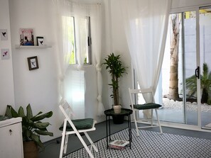 A room with a white easel, two chairs, a potted plant, a magazine on the floor, and a large window with sheer curtains. - Casa MARSI&ONDA -GARDEN FLOOR (Sámara)
