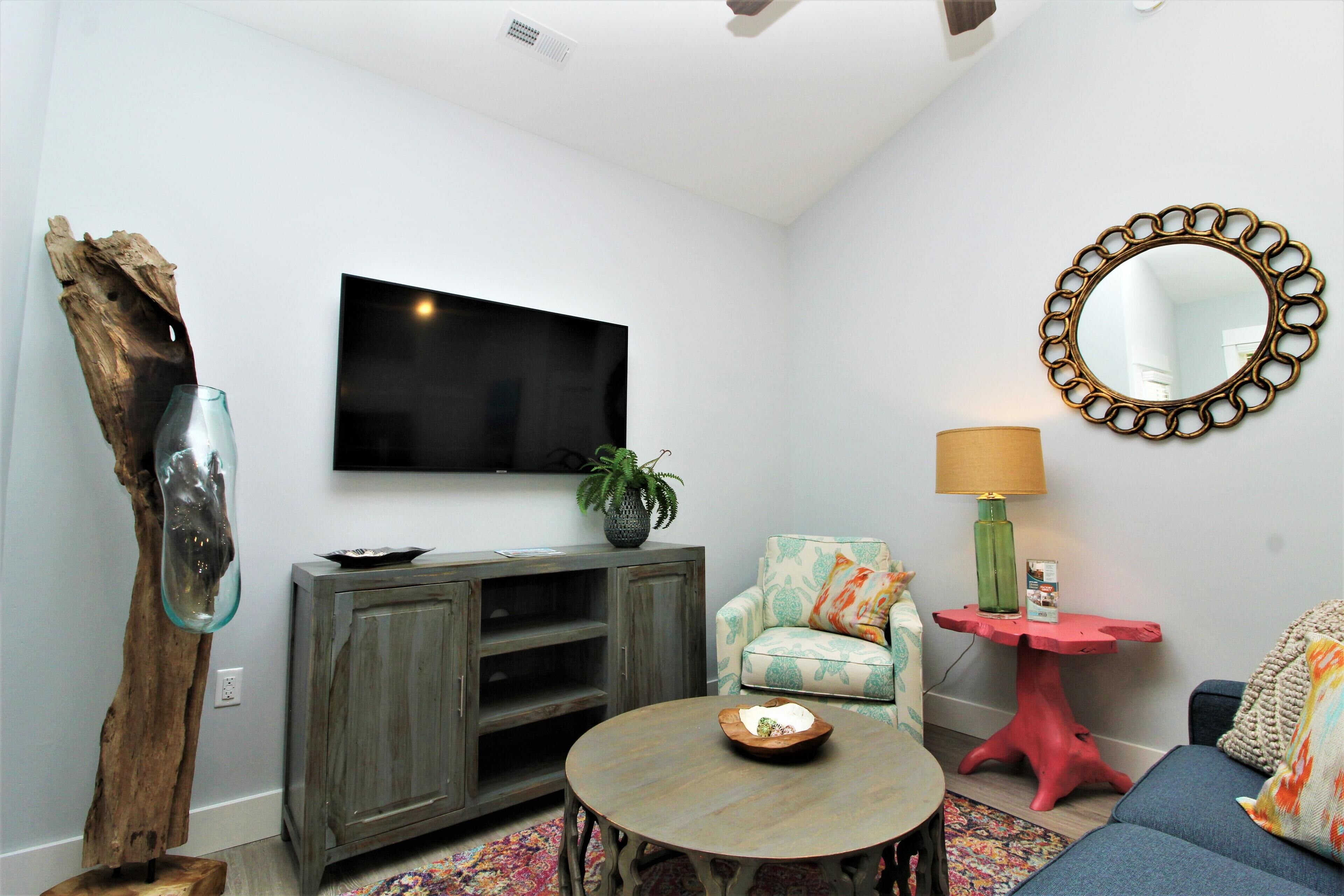 A living room with a flat-screen TV, a wooden cabinet, a round coffee table, a sofa, a red side table, and a mirror on the wall.