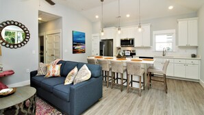 A modern kitchen with white cabinets, a dining area with bar stools, and a blue sofa in the living room. - Beacon Quarters by KEES Vacations (Corolla)