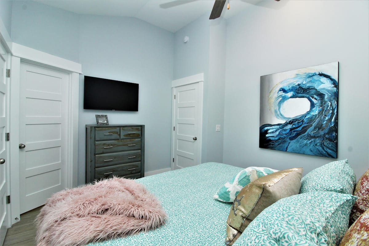 A bedroom with a bed, a fluffy pink ottoman, a dresser, and a painting on the wall.