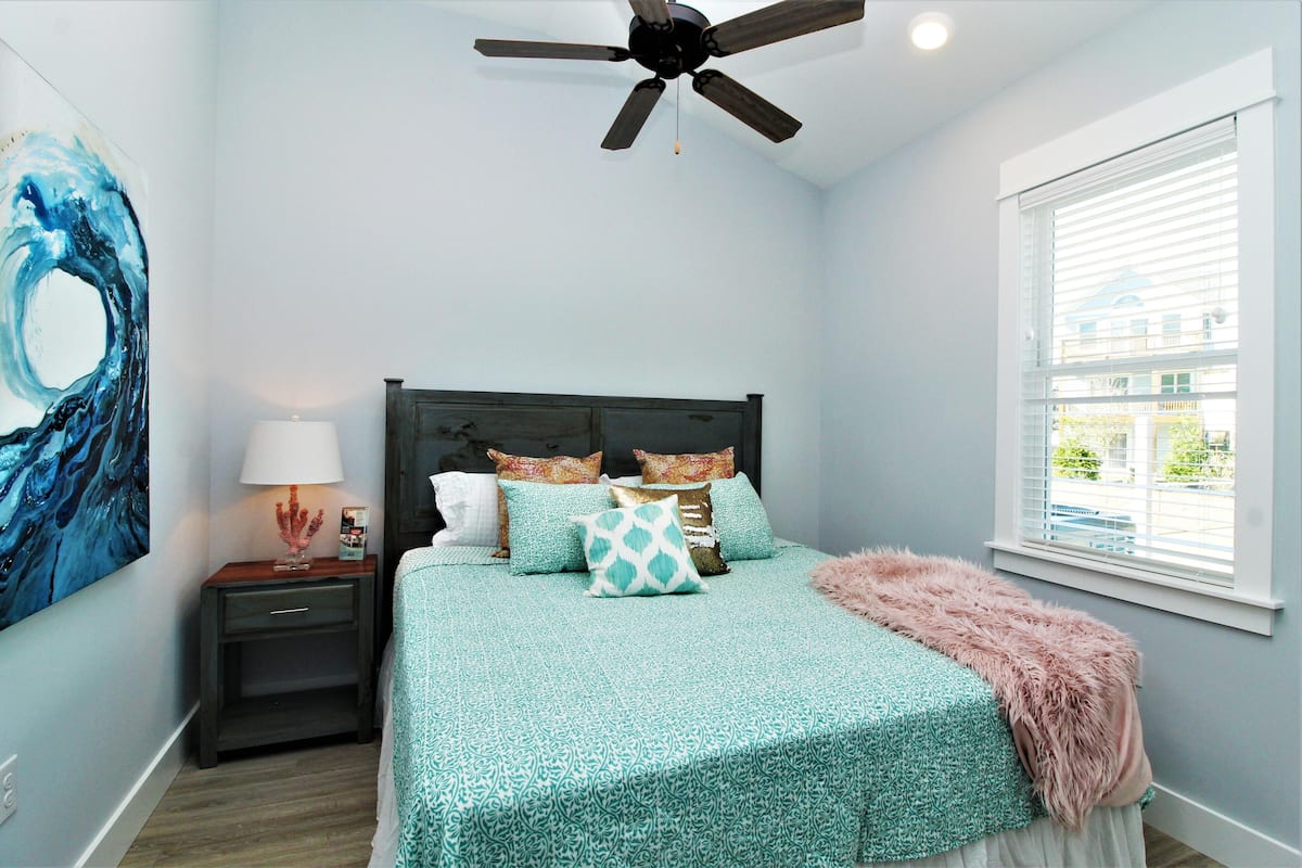 A bedroom with a bed, a nightstand, a lamp, a ceiling fan, and a window with blinds.