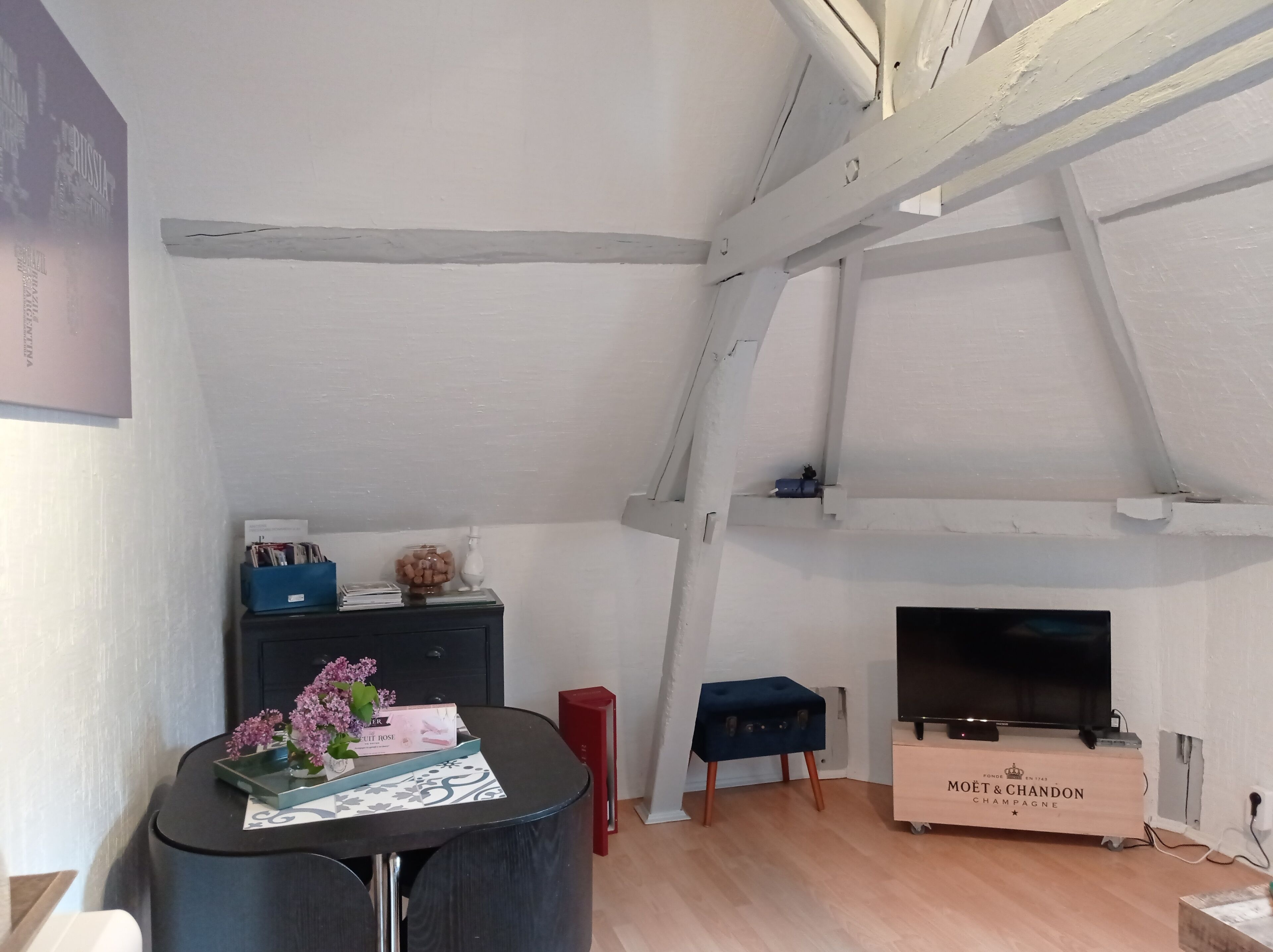 A loft-style room with exposed beams, a television, a small table with a vase of flowers, and a wooden cabinet labeled 'MOËT & CHANDON CHAMPAGNE.'