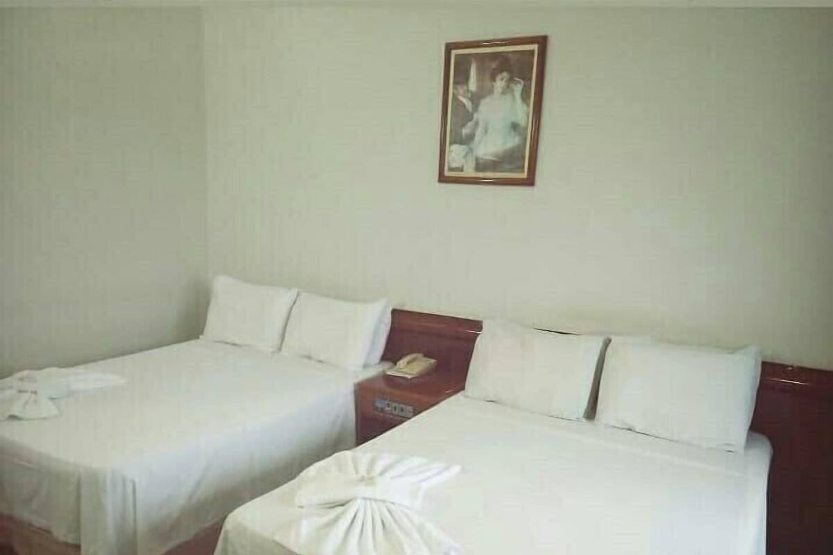 A hotel room with two single beds, each with a white bedspread and pillows, a wooden nightstand with a telephone, and a framed picture on the wall.