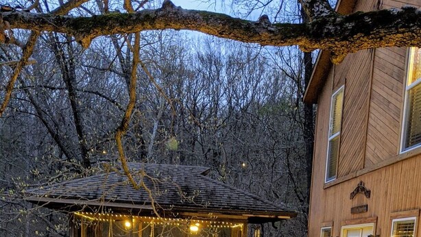 Eine Holzterrasse mit einer Steinsäule, eine kleine Sitzgelegenheit im Freien mit Lichterketten und ein Gebäude mit Holzfassade im Hintergrund.