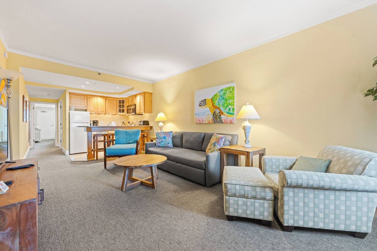 Ein modernes Wohnzimmer mit grauem Sofa, blauem Sessel und einem gemusterten Sessel. Ein Couchtisch und eine Lampe auf einem Beistelltisch sind ebenfalls zu sehen. Im Hintergrund ist eine Küche mit Holzschränken und einem weißen Kühlschrank erkennbar.