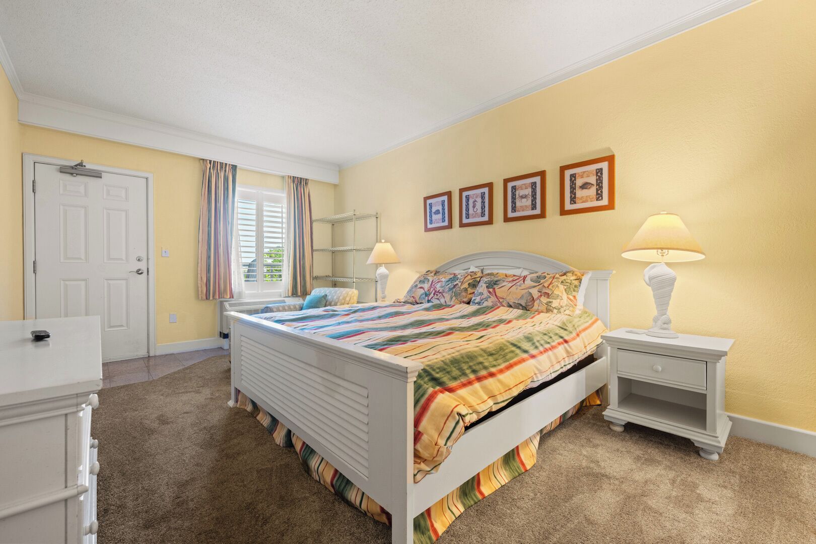 Une chambre à coucher comprenant un lit, des tables de chevet, une lampe, une commode, une porte et une fenêtre avec des rideaux.
