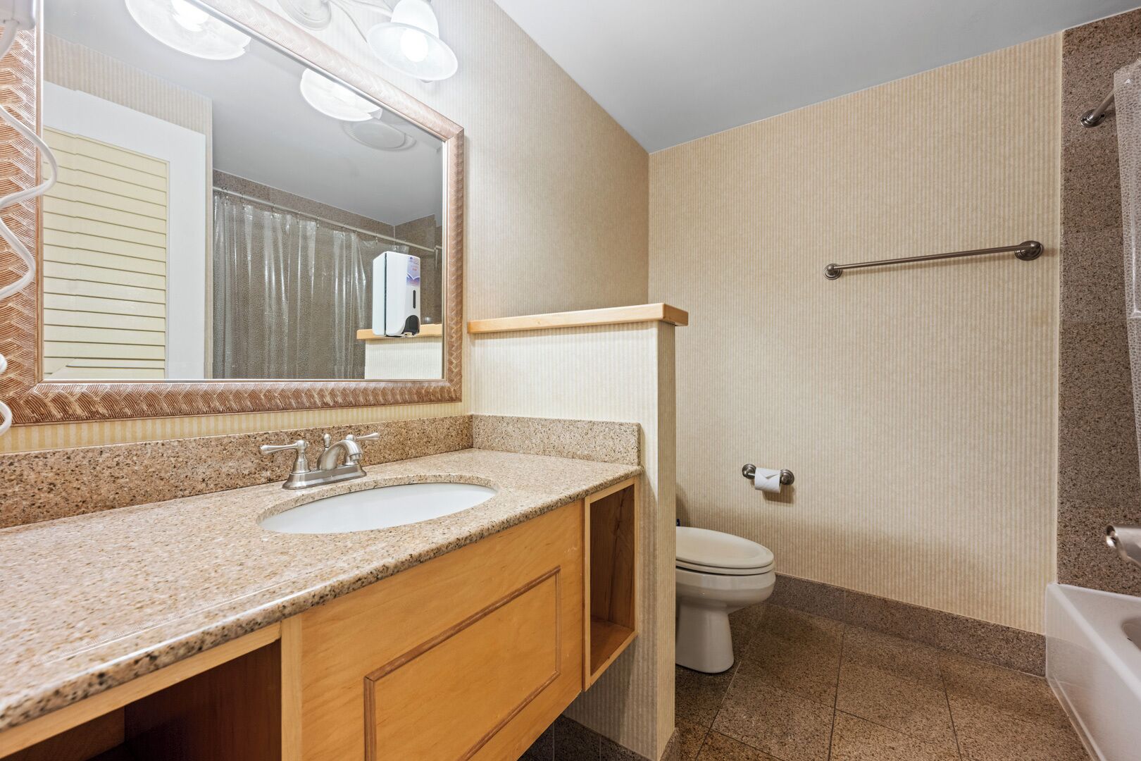 A bathroom with a granite countertop sink, a mirror with lights, a toilet, and a shower area.