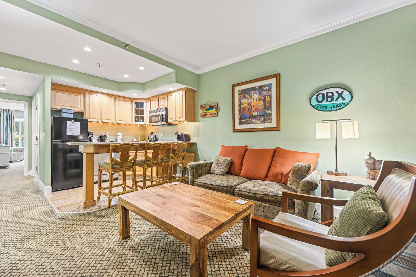 A cozy living room with a wooden coffee table, a sofa with plaid cushions, and a kitchen area with wooden cabinets and a refrigerator.