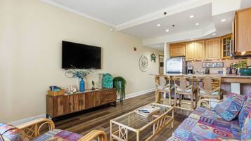 A living room with a sofa, chairs, a coffee table, a TV, a kitchen area with wooden cabinets, and a dining table.
