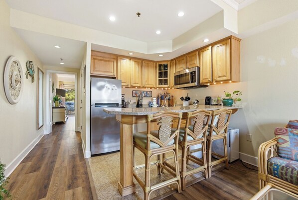 A kitchen with wooden cabinets, a stainless steel refrigerator, a microwave, and a bar area with wicker chairs.