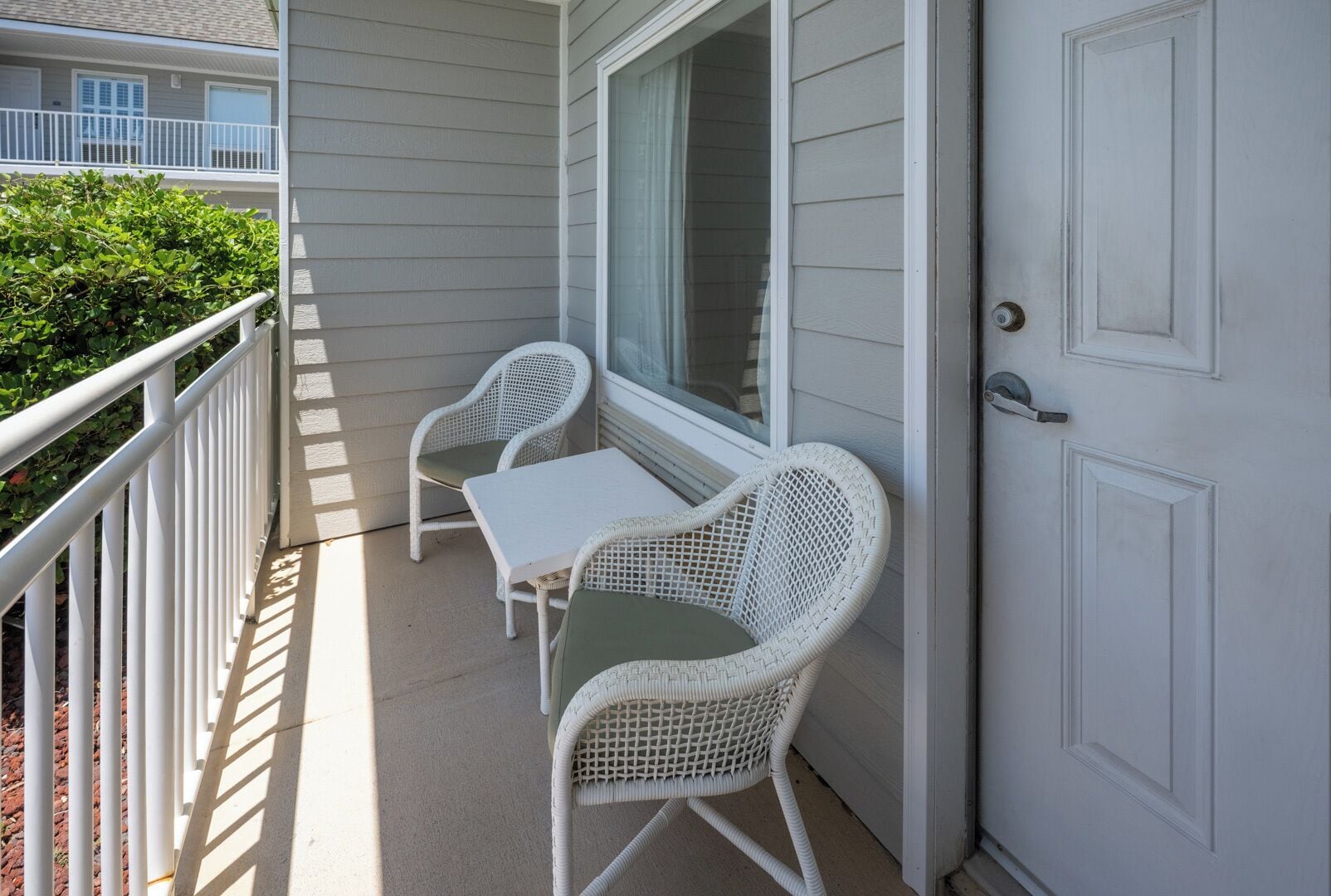 A balcony with two white wicker chairs and a small table, a white door, and a white railing.