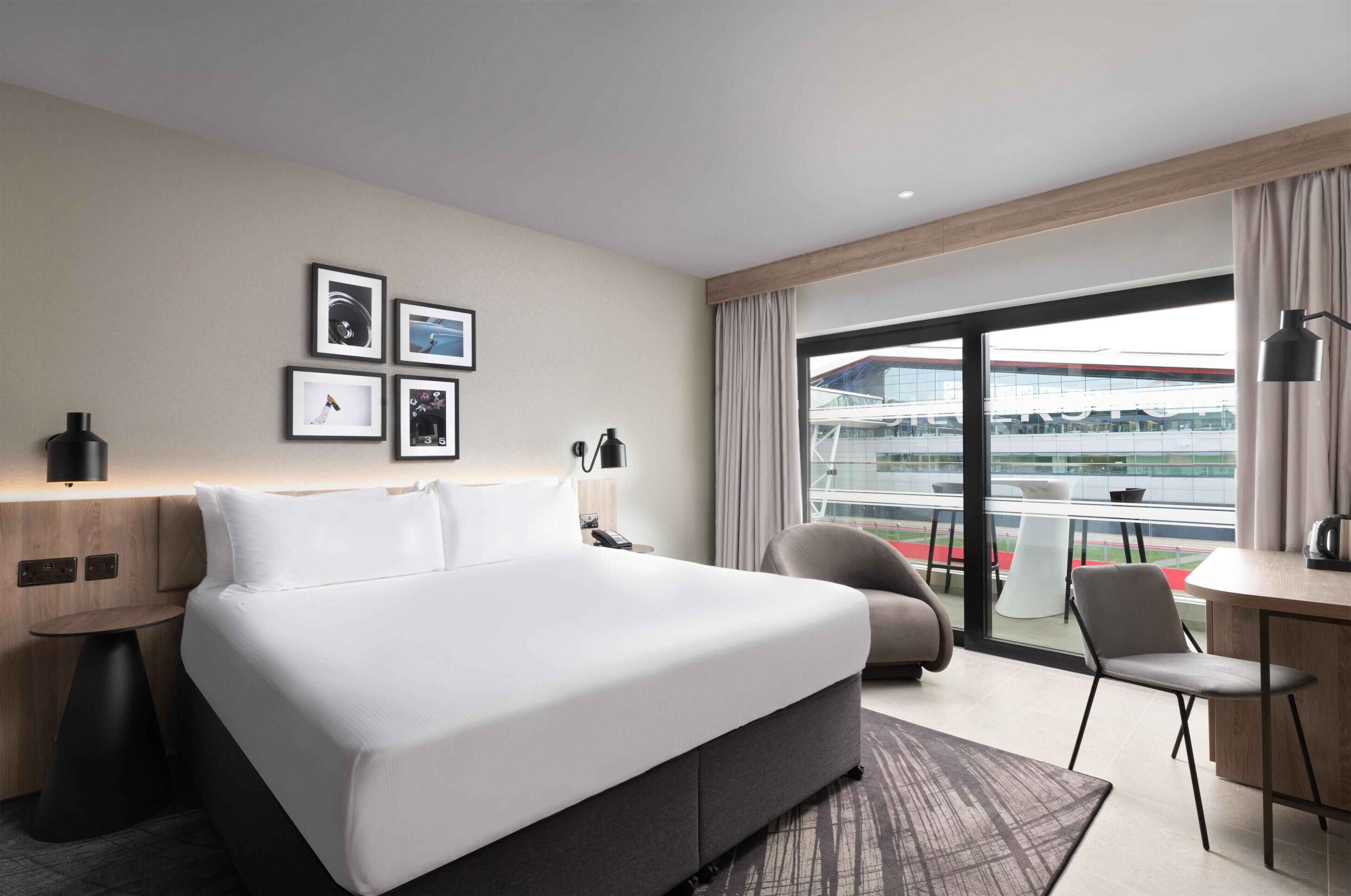 Ein Hotelzimmer mit einem großen Bett, einem Schreibtisch, einem Stuhl, einer Lampe und Blick auf ein Gebäude durch das Fenster.