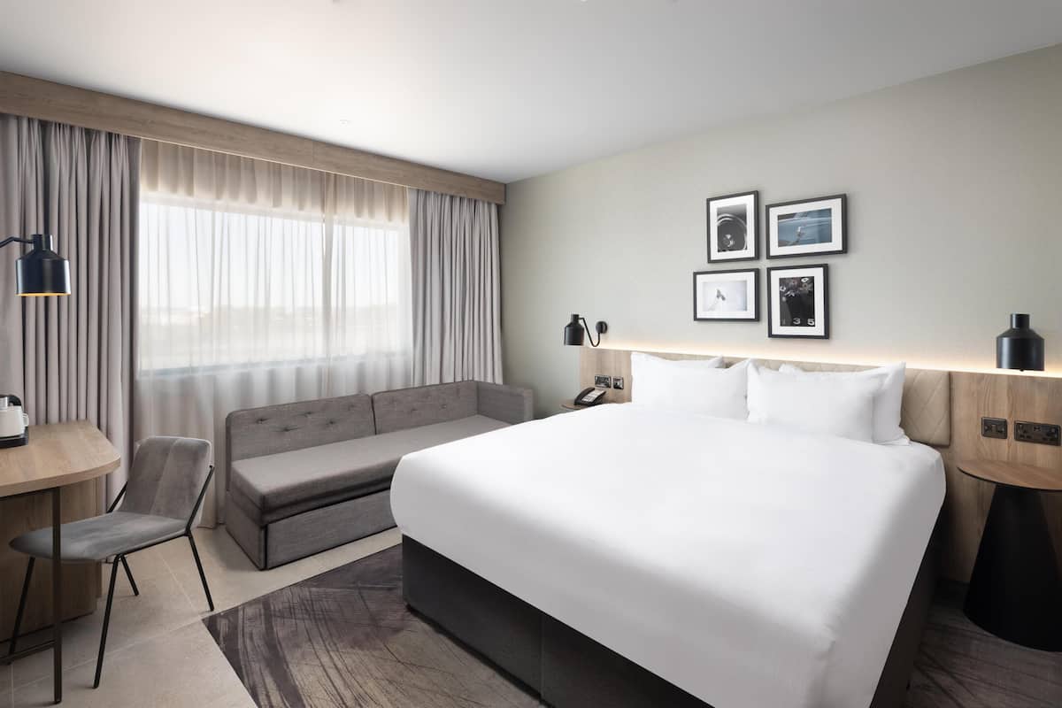 Ein Hotelzimmer mit einem großen Bett, einem Sofa, einem Schreibtisch mit Stuhl und einem Fenster mit Vorhängen.