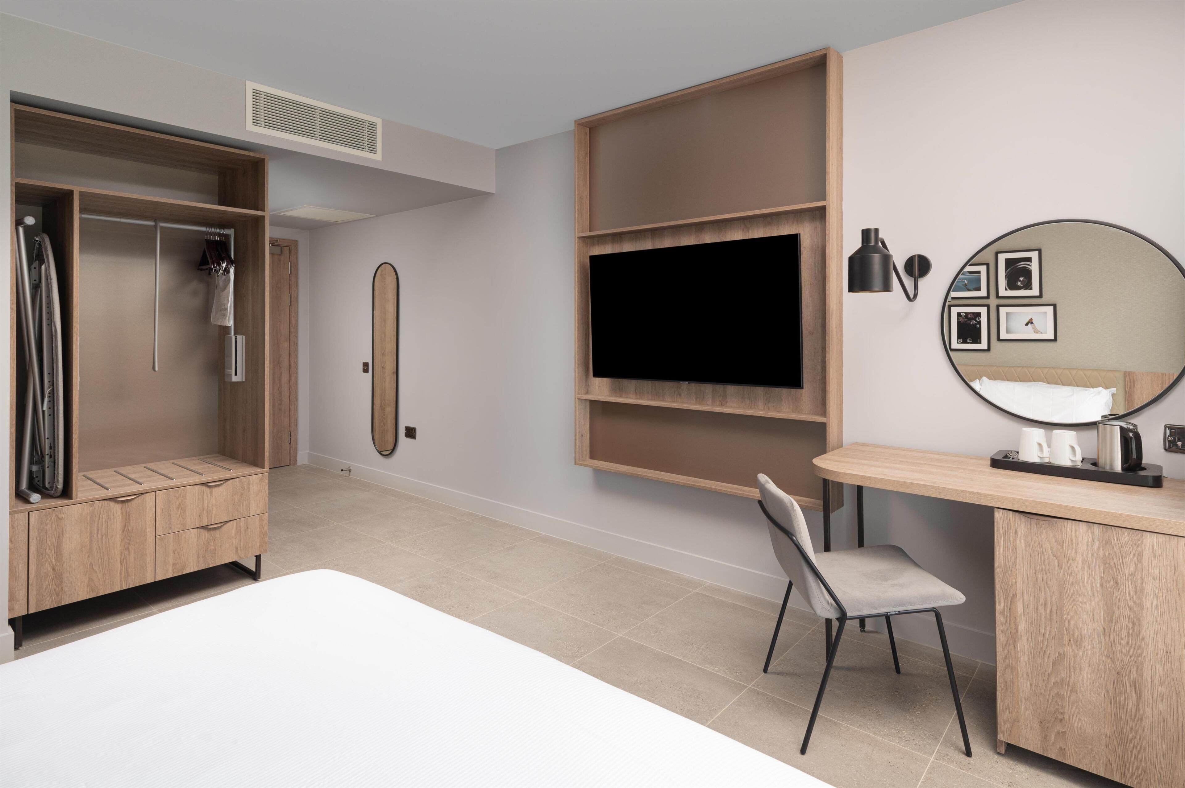 A modern hotel room with a bed, a desk with a chair, a wardrobe, a TV, and a wall-mounted shelf.