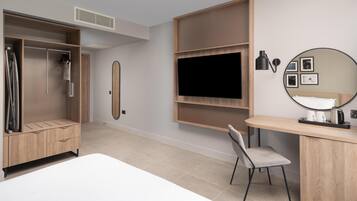 A modern hotel room with a bed, a desk with a chair, a wardrobe, a TV, and a wall-mounted shelf.