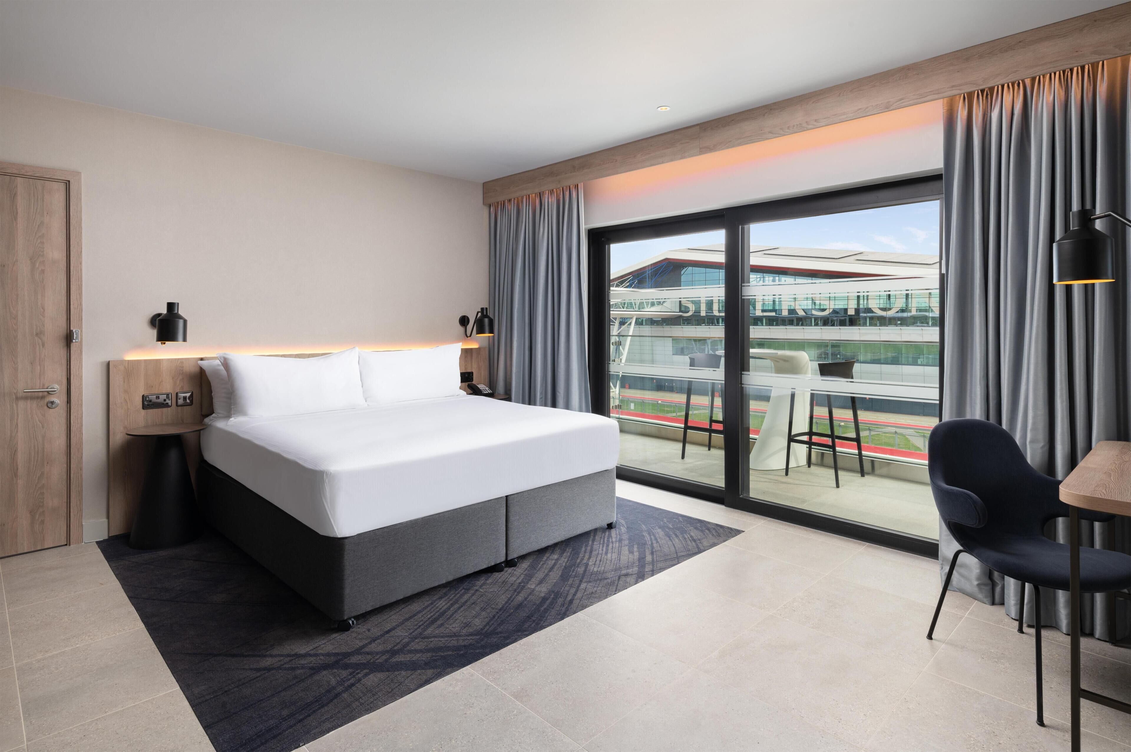 A modern hotel room with a large bed, a desk, and a view of a stadium.