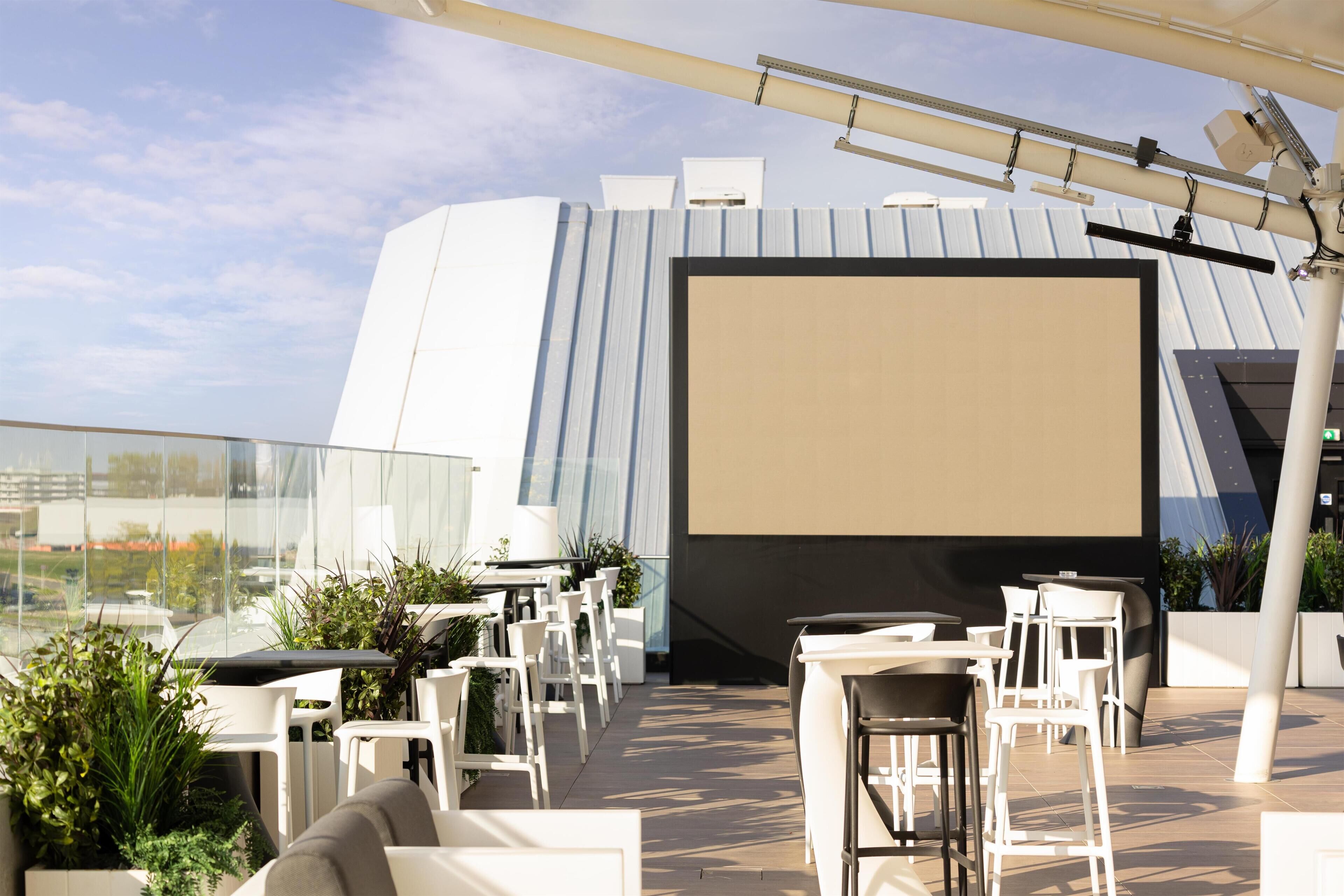 An outdoor seating area with tables and chairs, a large screen, and potted plants.