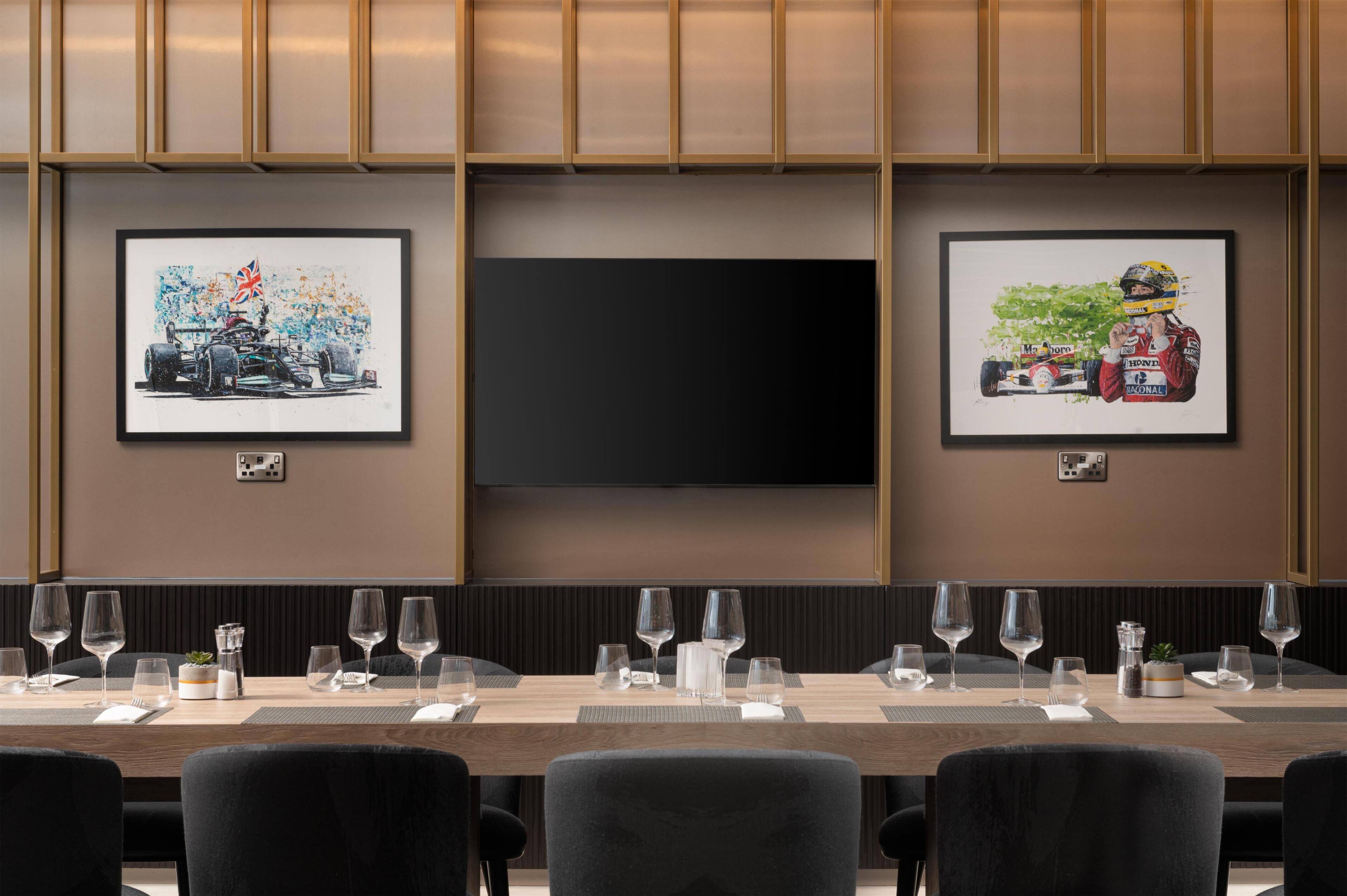 A conference room with a long table set for a meeting, framed artwork on the walls, and a flat-screen TV.