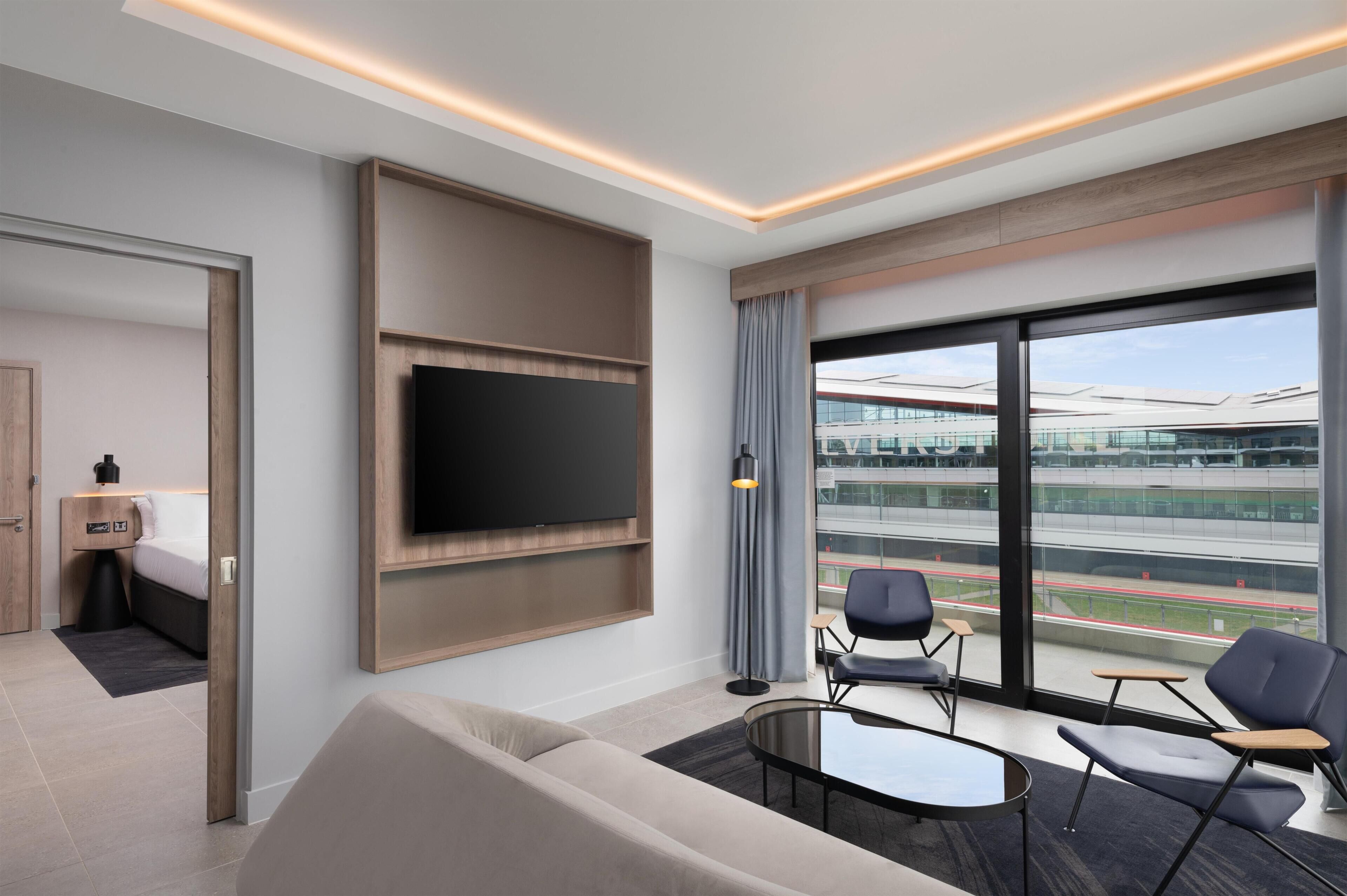 Een moderne hotelkamer met een grote televisie, comfortabele zitplaatsen en een balkon met uitzicht op een stadion.
