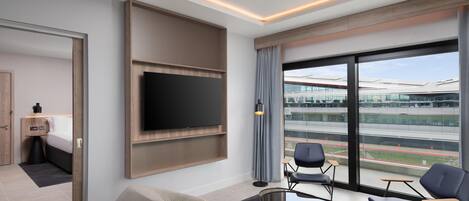 A modern hotel room with a large TV, comfortable seating, a balcony with a view of a stadium.
