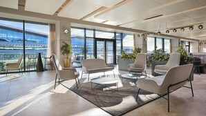 A modern hotel lobby with large windows, seating areas, and a view of a stadium. - Hilton Garden Inn Silverstone (Towcester)