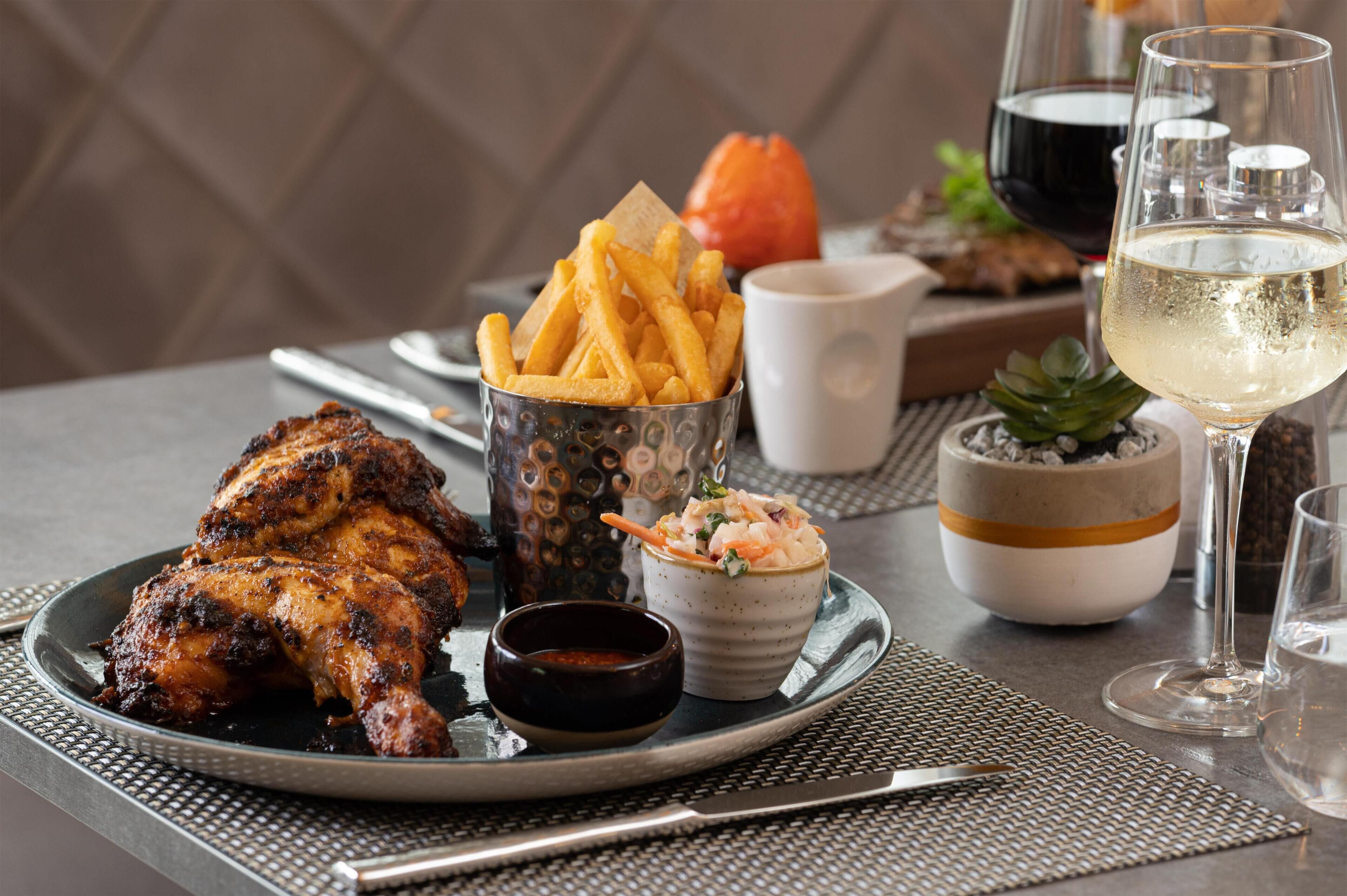A dining table with a plate of grilled chicken, French fries, a small bowl of salad, a glass of white wine, and a glass of water.