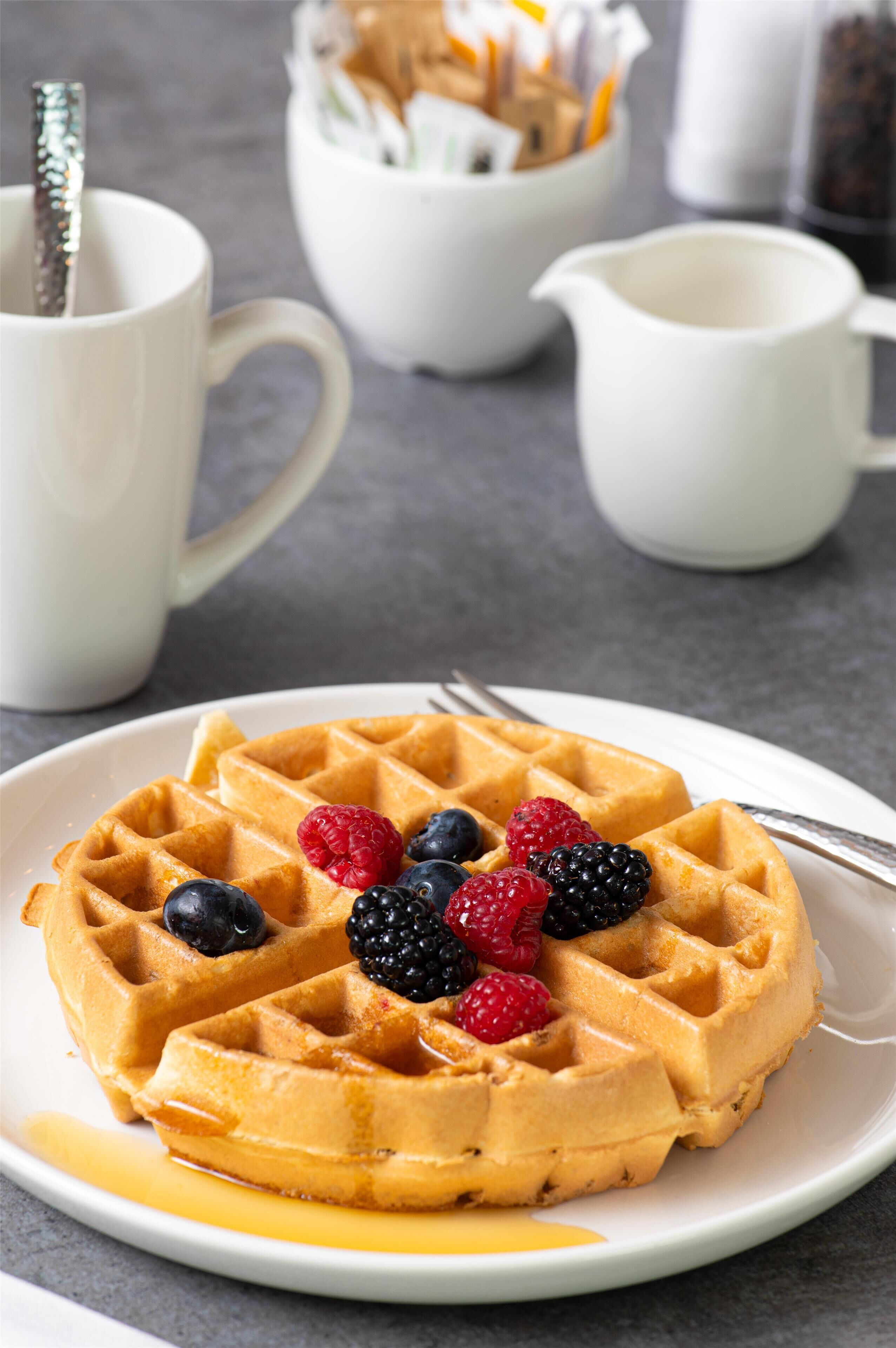 Une assiette de gaufres garnies de fruits rouges et nappées de sirop, une fourchette, et une tasse blanche en arrière-plan.