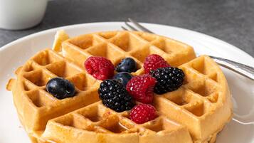 Une assiette de gaufres garnies de fruits rouges et nappées de sirop, une fourchette, et une tasse blanche en arrière-plan.