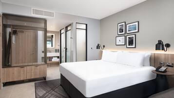 Una habitación de hotel moderna con una cama grande, un armario de madera, un baño con separador de vidrio y una pared decorada con cuadros.