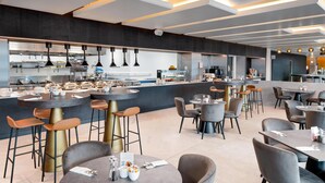 A modern restaurant with a central bar area, multiple seating options, and a visible kitchen in the background. - Hilton Garden Inn Silverstone (Towcester)
