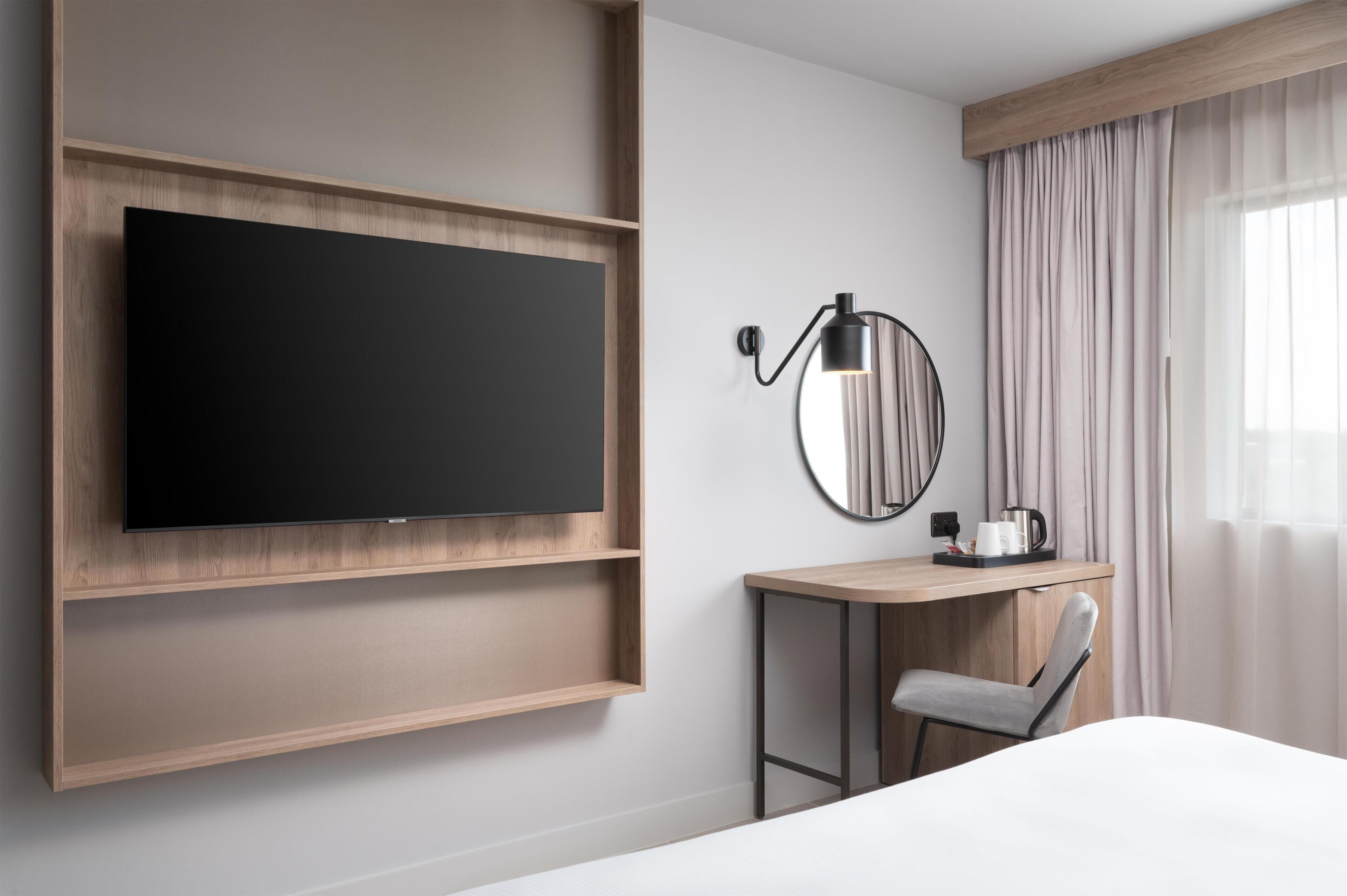 Ett modernt hotellrum med en väggmonterad TV, ett skrivbord med stol, en kaffebryggare och ett stort fönster med gardiner.