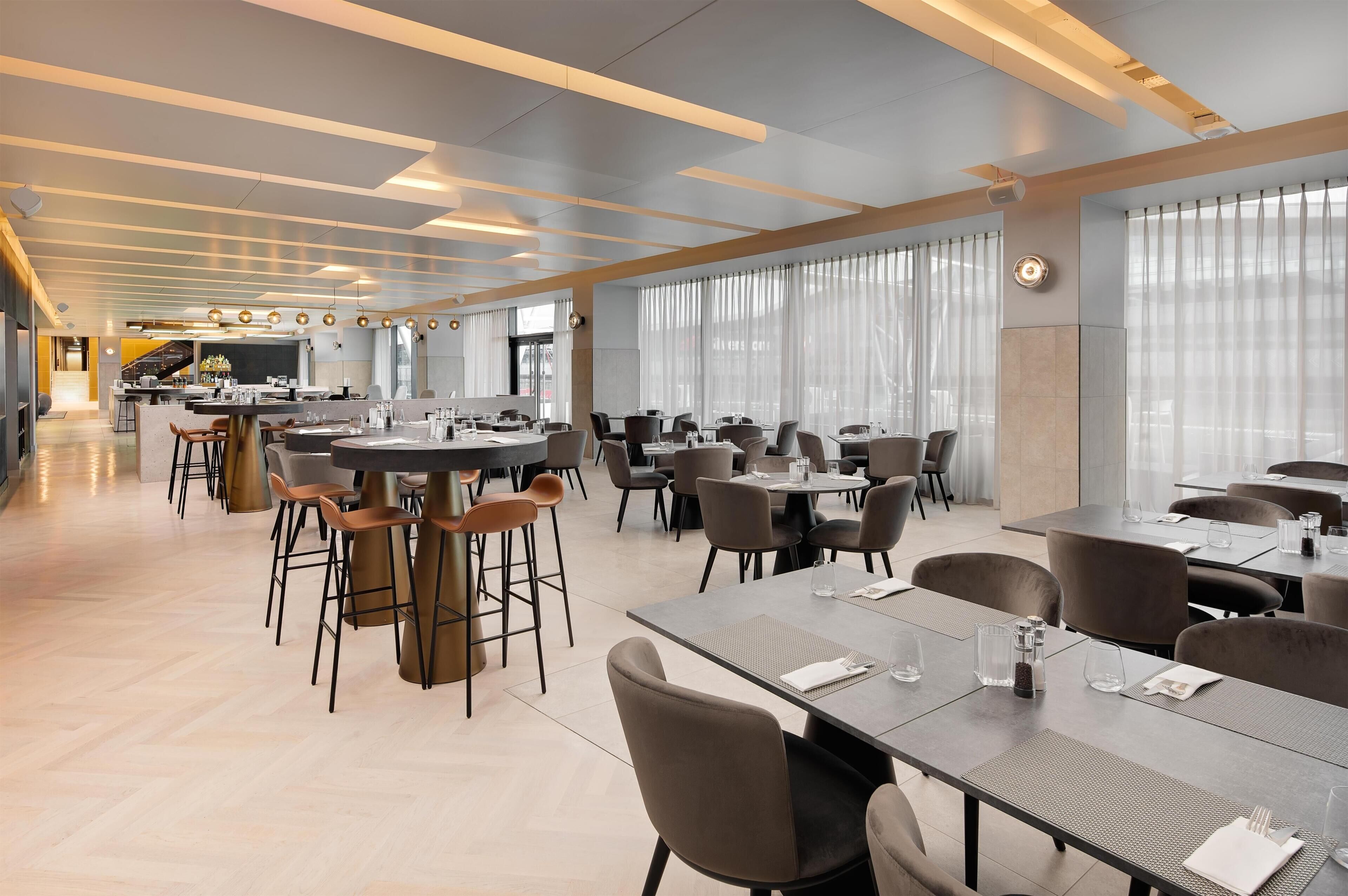 A modern dining area with tables and chairs, a kitchen in the background, and large windows.