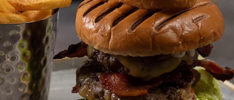 Una hamburguesa con pan, lechuga, tomate, queso y tocino, acompañada de papas fritas y una taza pequeña.