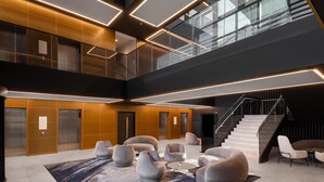 A modern lobby with a central seating area, a staircase, and a mezzanine level with glass railings. - Hilton Garden Inn Silverstone (Towcester)