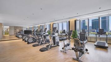 A modern gym with various cardio machines, large windows offering city views, and a wooden floor.