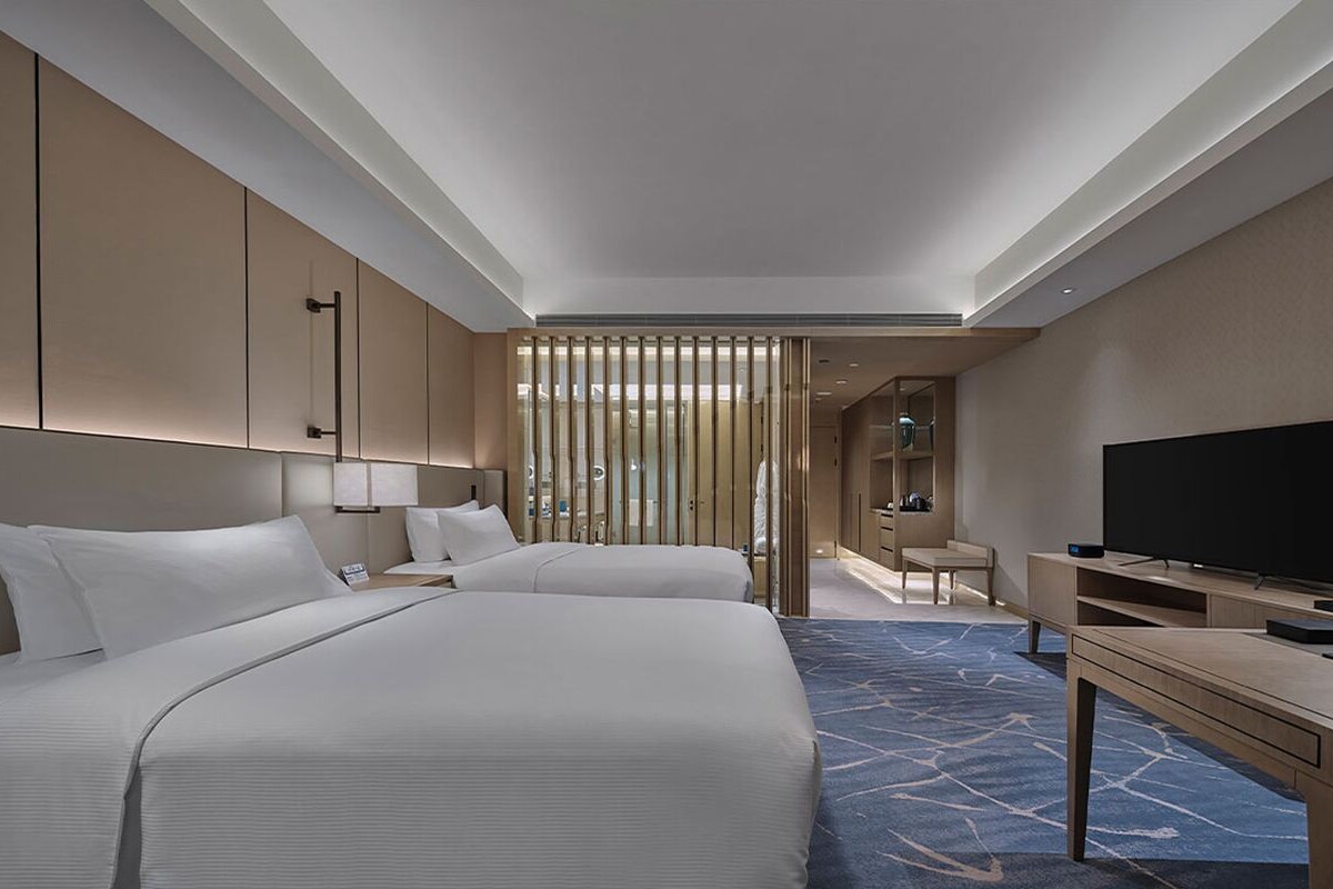 A hotel room with two beds, a TV, a desk, and a bathroom visible through a partition.