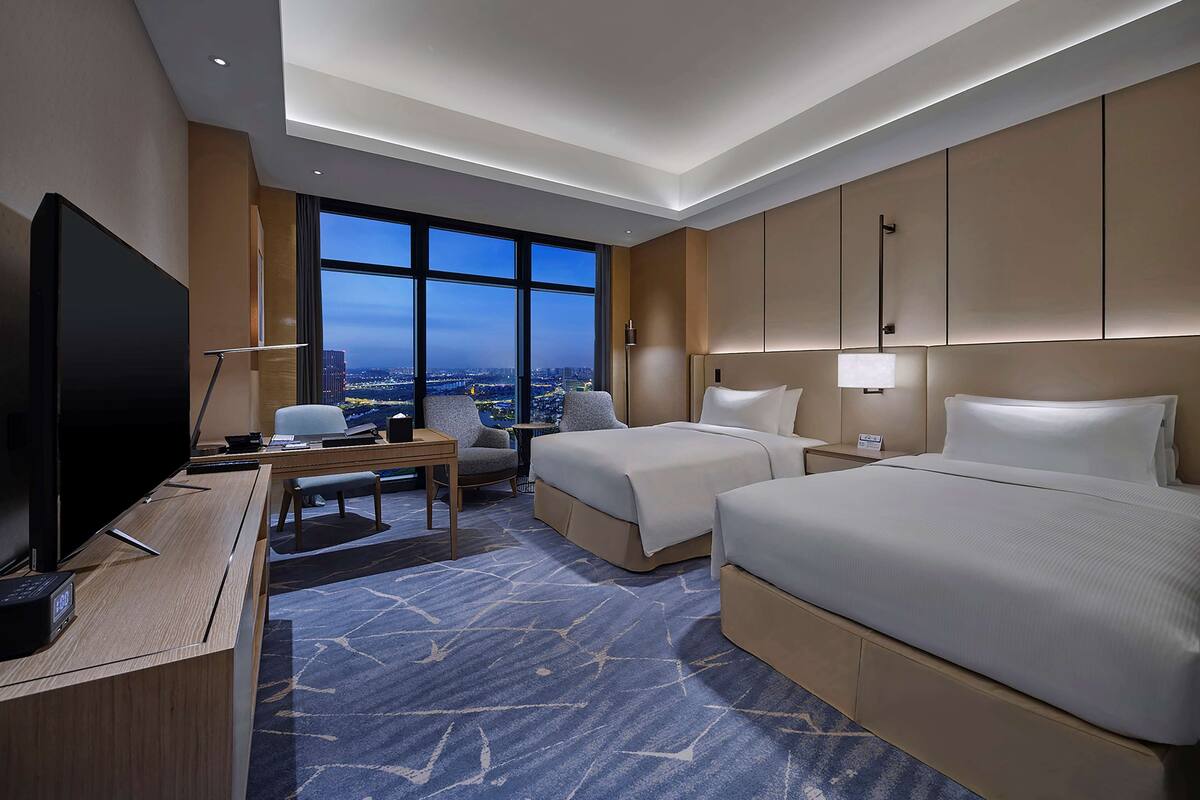 Habitación de hotel con dos camas, un escritorio con computadora, televisión y un ventanal con vista a la ciudad.