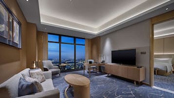 A modern hotel room with a large window offering a city view, a flat-screen TV, a sofa, a coffee table, and a bed with a headboard.