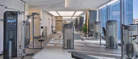 A modern gym with various exercise machines, a bench, and large windows offering views of the city.