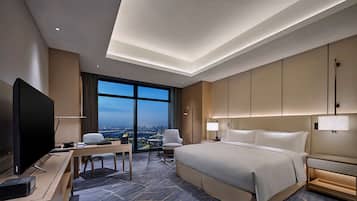 A modern hotel room with a large bed, a desk with a flat-screen TV, a chair, and a view of the cityscape through large windows.