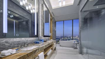 A modern bathroom with a large mirror, a marble countertop, a bathtub, and a city view through large windows.