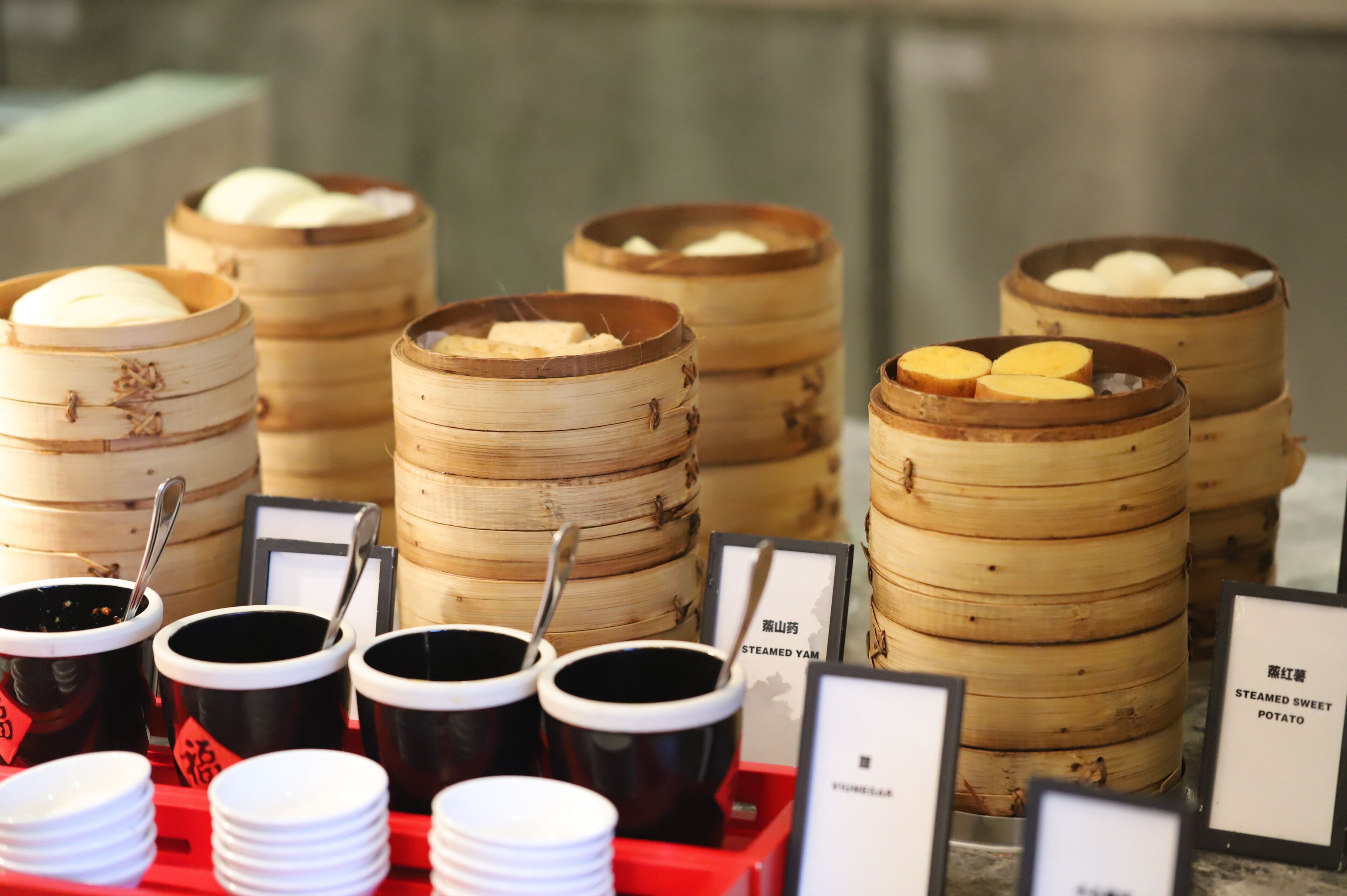Several bamboo steamers with food, accompanied by black and white cups and plates, likely for a buffet or food service.