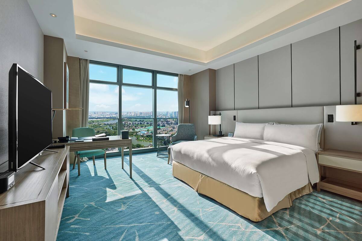 Una habitación de hotel moderna con una cama grande, un escritorio, un televisor y vistas de la ciudad a través de amplias ventanas.