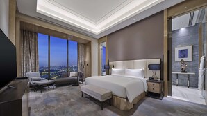 A modern hotel room with a large bed, a seating area with a sofa and armchair, a desk, and a bathroom with a glass shower. - Hilton Beijing Tongzhou (Tongzhou)