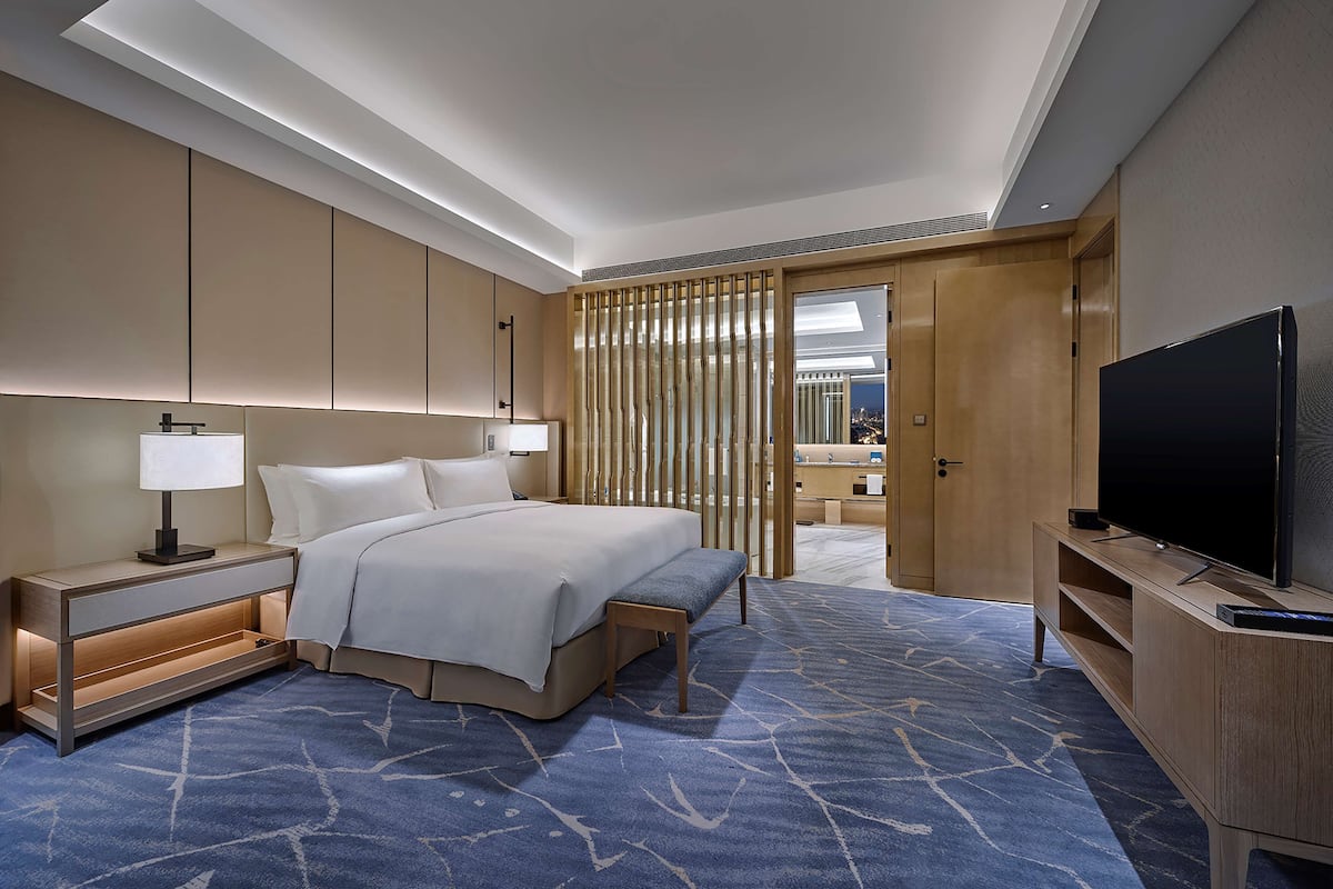 Habitación de hotel moderna con una cama grande, mesitas de noche, televisión y vistas de la ciudad a través de una puerta corredera.