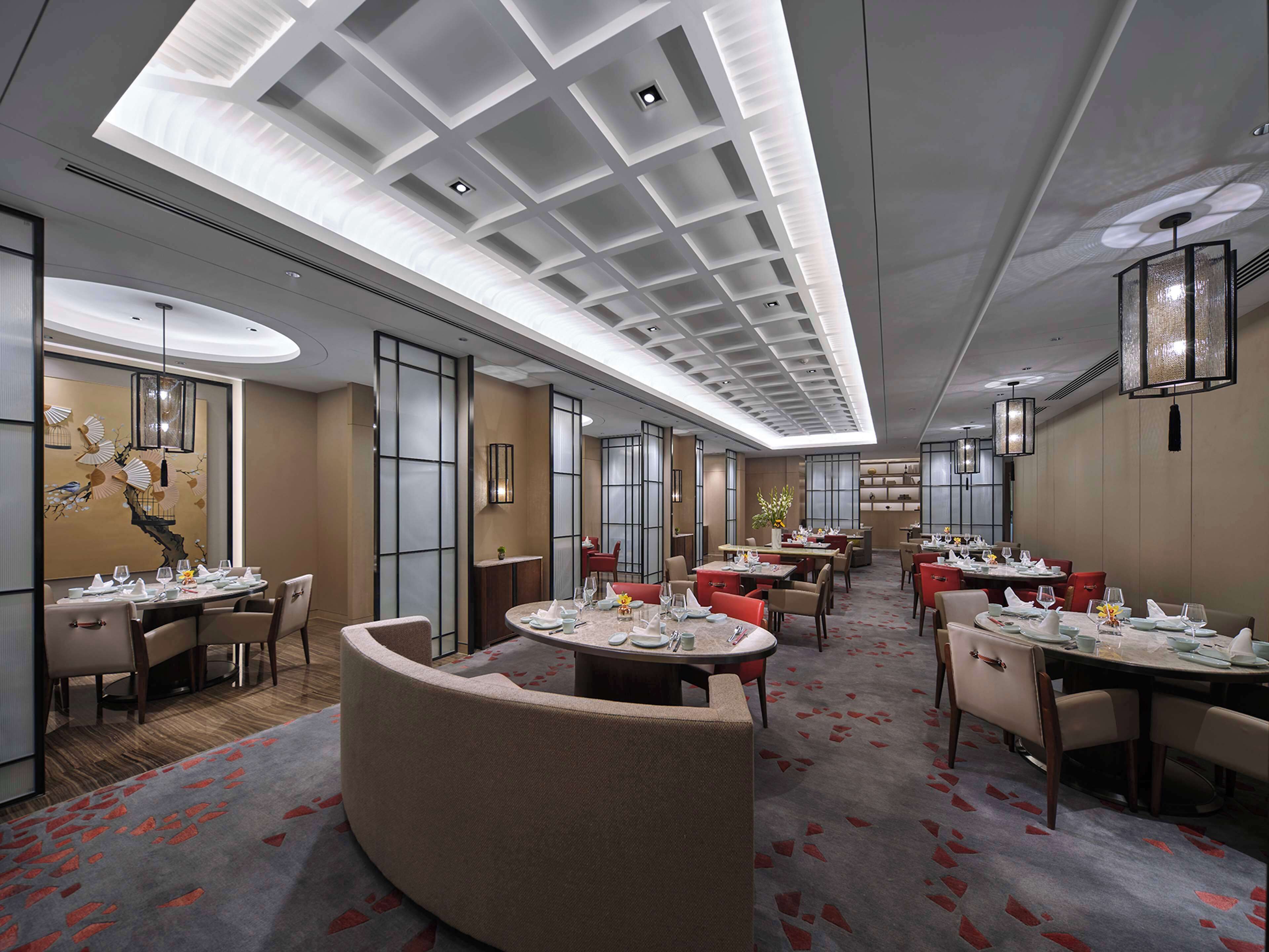 A modern hotel dining area with multiple tables and chairs, a central seating area, and a ceiling with recessed lighting.