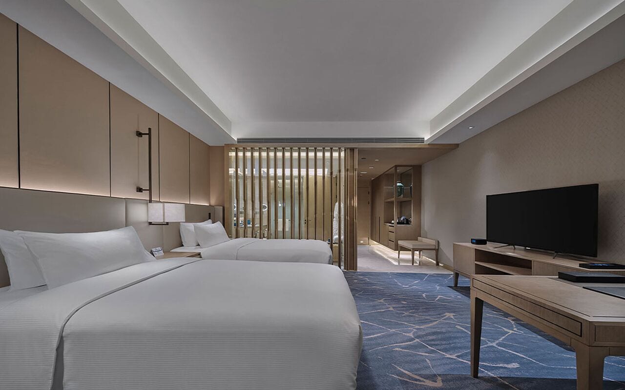 A hotel room with two beds, a TV, a desk, and a bathroom visible through a partition.